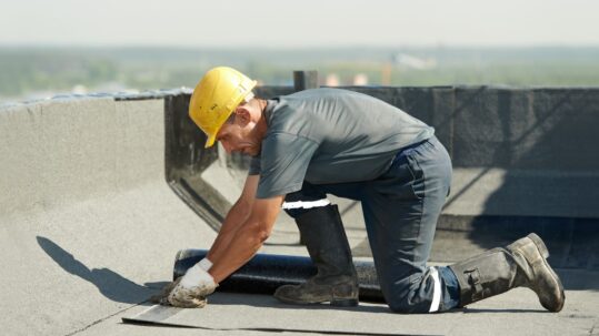 Worker in a yellow hard hat repairing a flat roof, kneeling and applying roofing material.