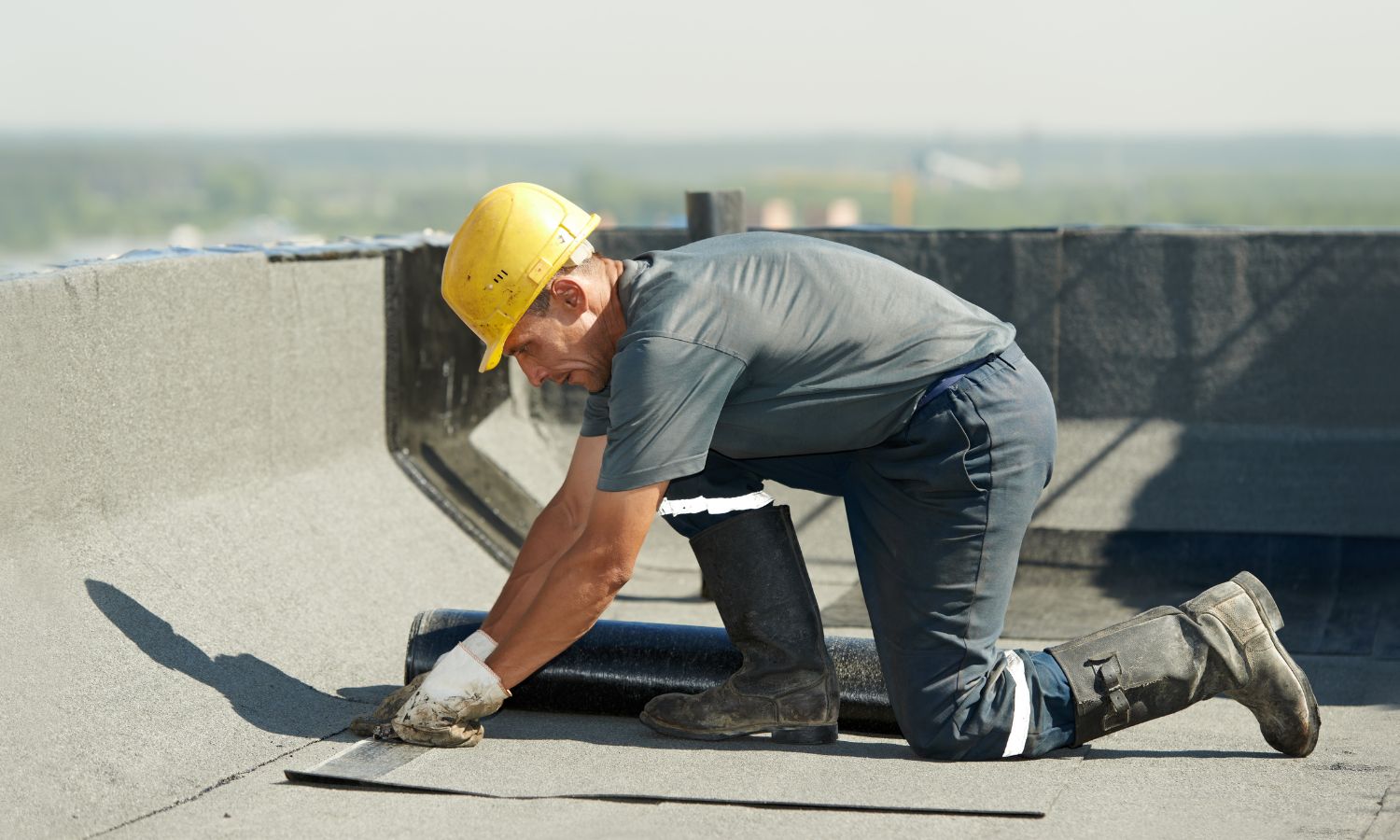 Worker in a yellow hard hat repairing a flat roof, kneeling and applying roofing material.